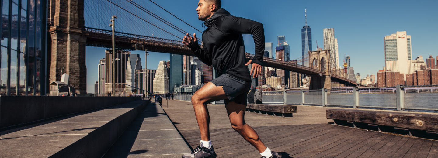 A man is running on a boardwalk near the Brooklyn Bridge with the New York City skyline in the background.